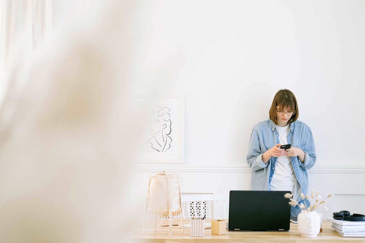 Woman in casual denim texting on smartphone in a bright, minimalist home office setup.